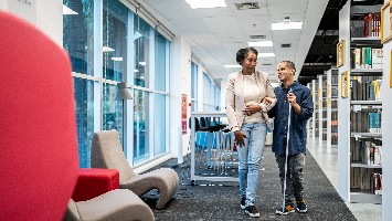 Woman helping man with cane walking in library.