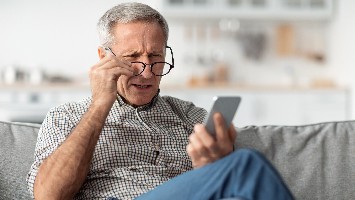 Senior man adjusts glasses and holds up phone.