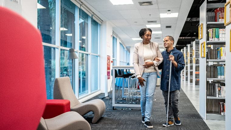 Woman helping man with cane walking in library.