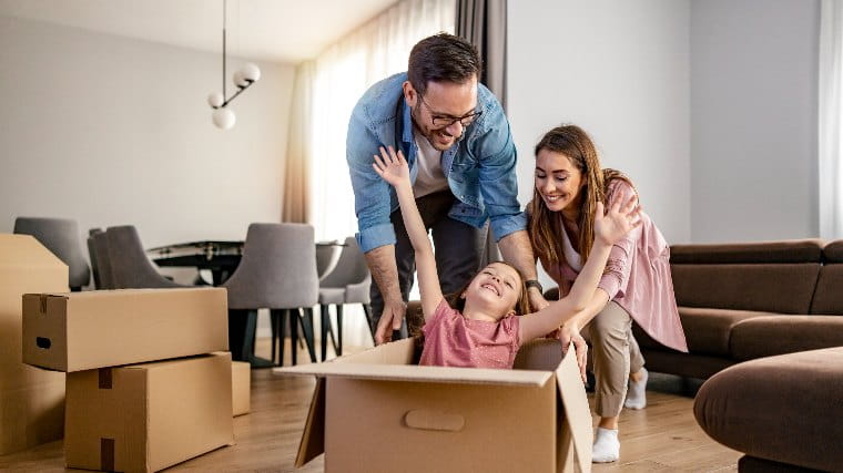 Parents playing with young daughter who is sitting in open moving box in new home.