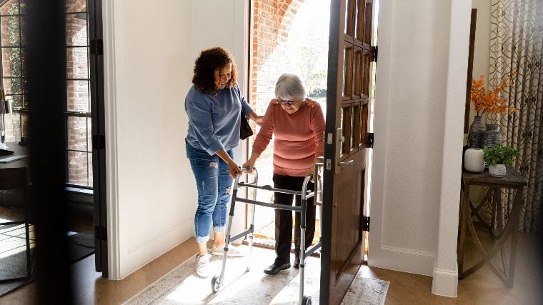 Caretaker helping senior woman with walker through doorway.