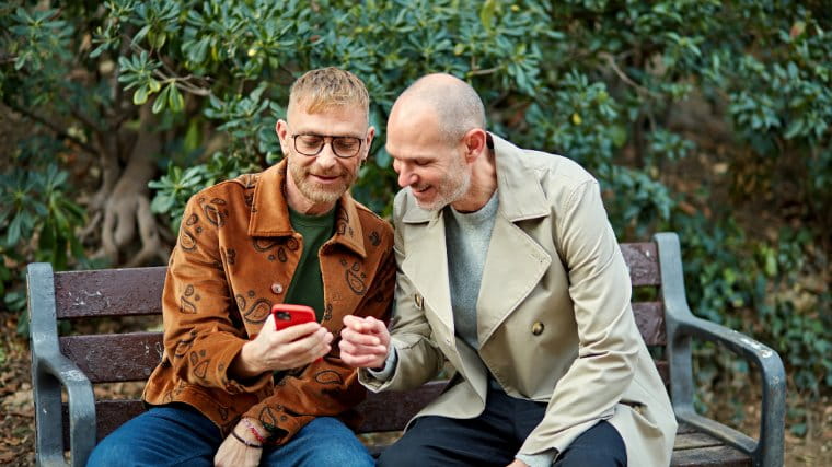 Two men sitting on a bench and looking at the same phone.