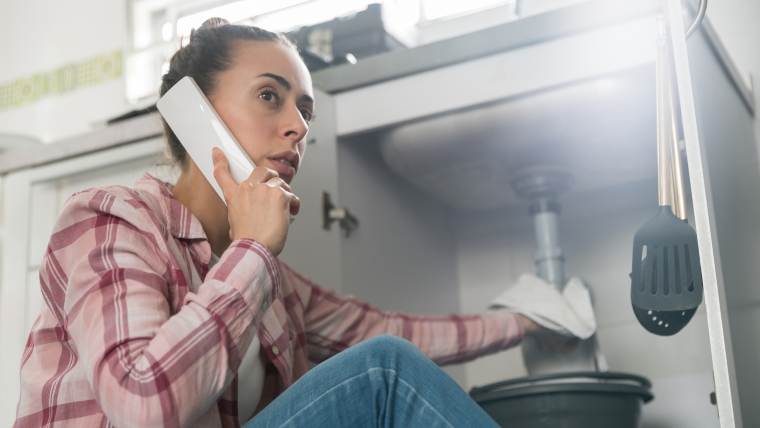 Woman trying to fix a pipe under the sink while on the phone with a plumber.