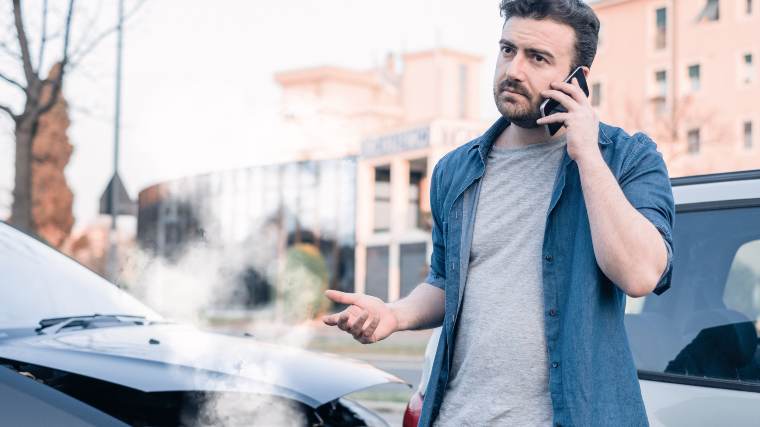 Man using cellphone to call for assistance, standing next to a car with smoke pouring out of the engine.