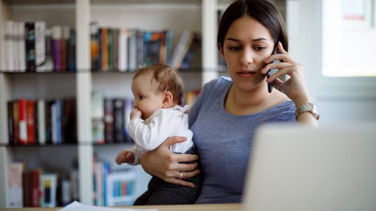 Mother holding small baby in one arm as she talks on the phone and reads from a laptop.
