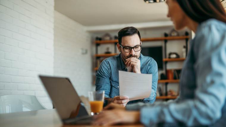 Man reviewing a mortgage bill while his wife works on a laptop in the foreground.