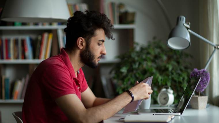 Young man reading a bill, sitting in front of a laptop, trying to decide if he should pay it.