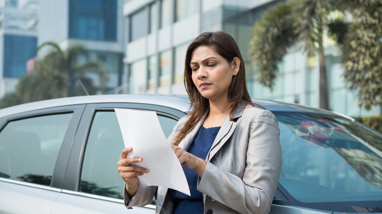 Woman reads paperwork while standing next to car.
