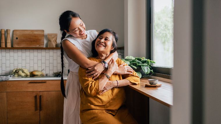 Mother and adult daughter embrace in the kitchen.