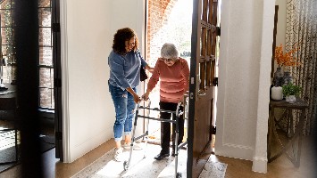 Caretaker helping senior woman with walker through doorway.
