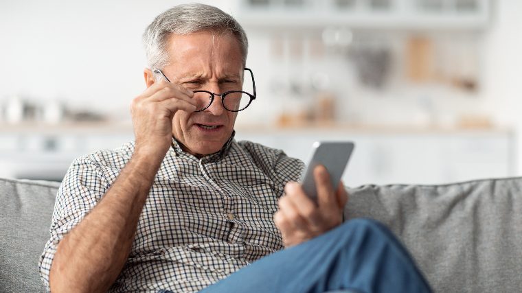 Senior man adjusts glasses while reading phone.