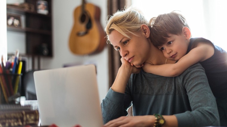 Stressed mother hugged by young son while looking at laptop.